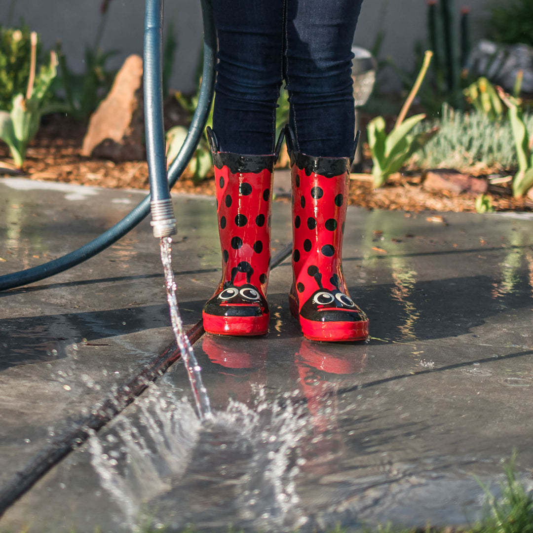 Close up of a kid's leg wearing Western Chief Kids Lucy Ladybug red rain boots outside holding a hose that pouring out water.