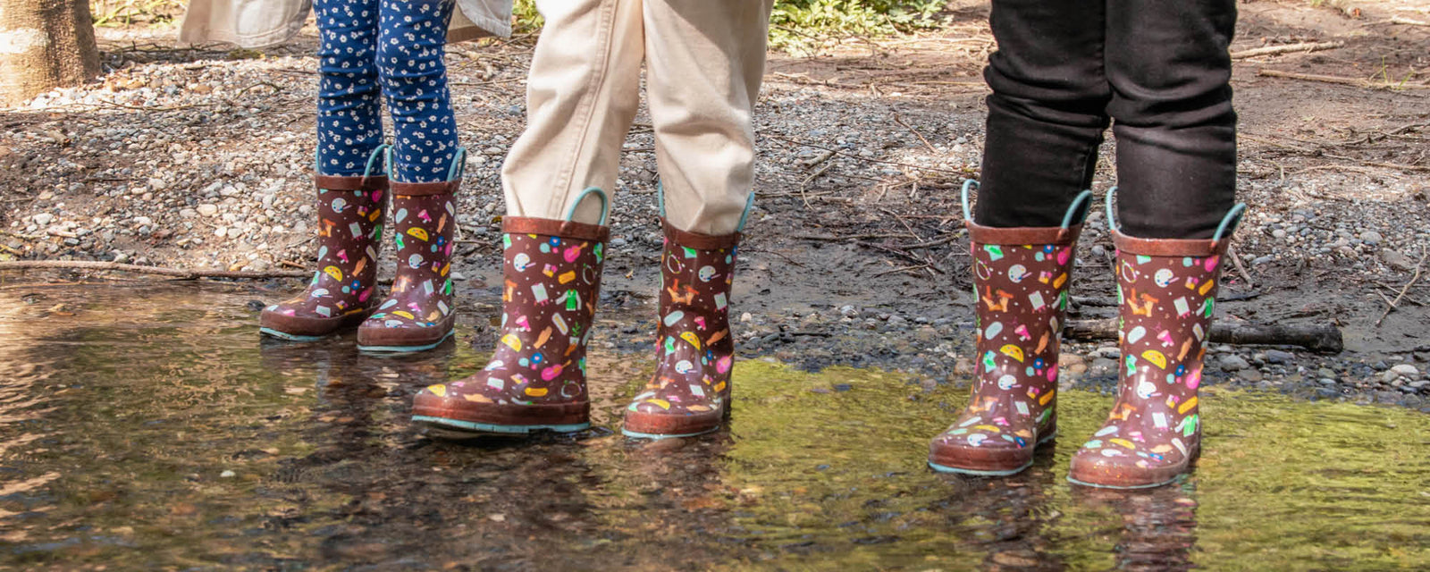 close up of three kids legs wearing Western Chief Girl Scouts themed Rain Boots standing in a puddle of water. 