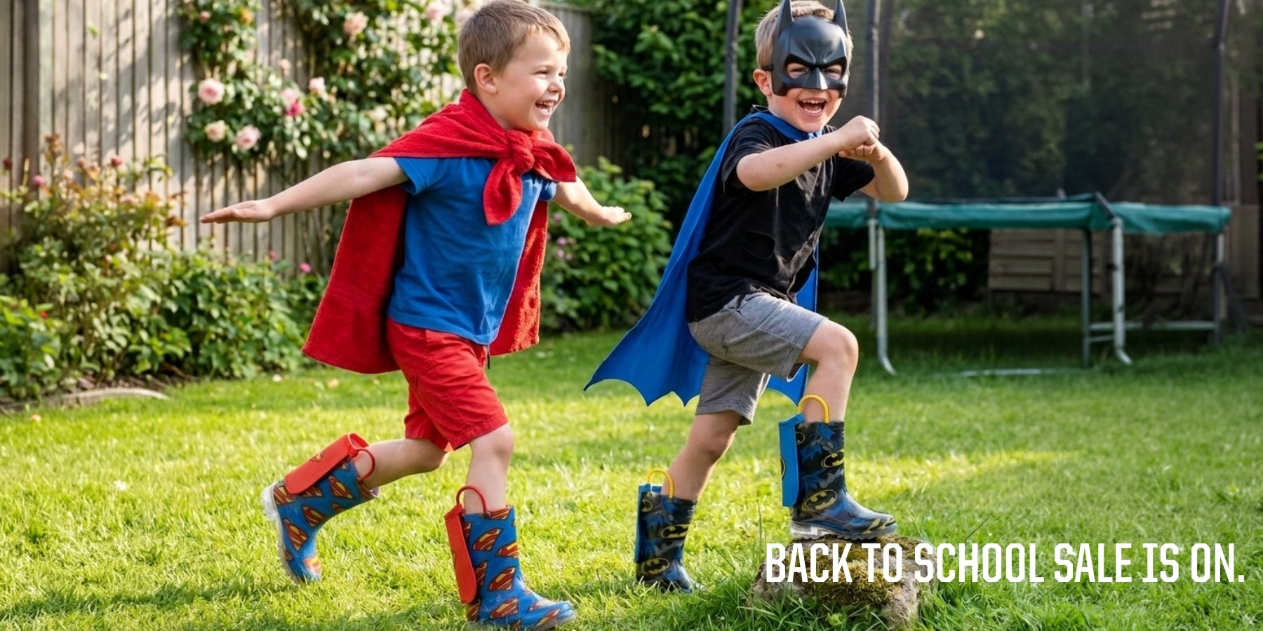 Two children running and playing in a backyard wearing superhero costumes. One child wears a blue and red Superman-style costume with a red cape, red shorts, and blue boots. contains-synthetic-performers