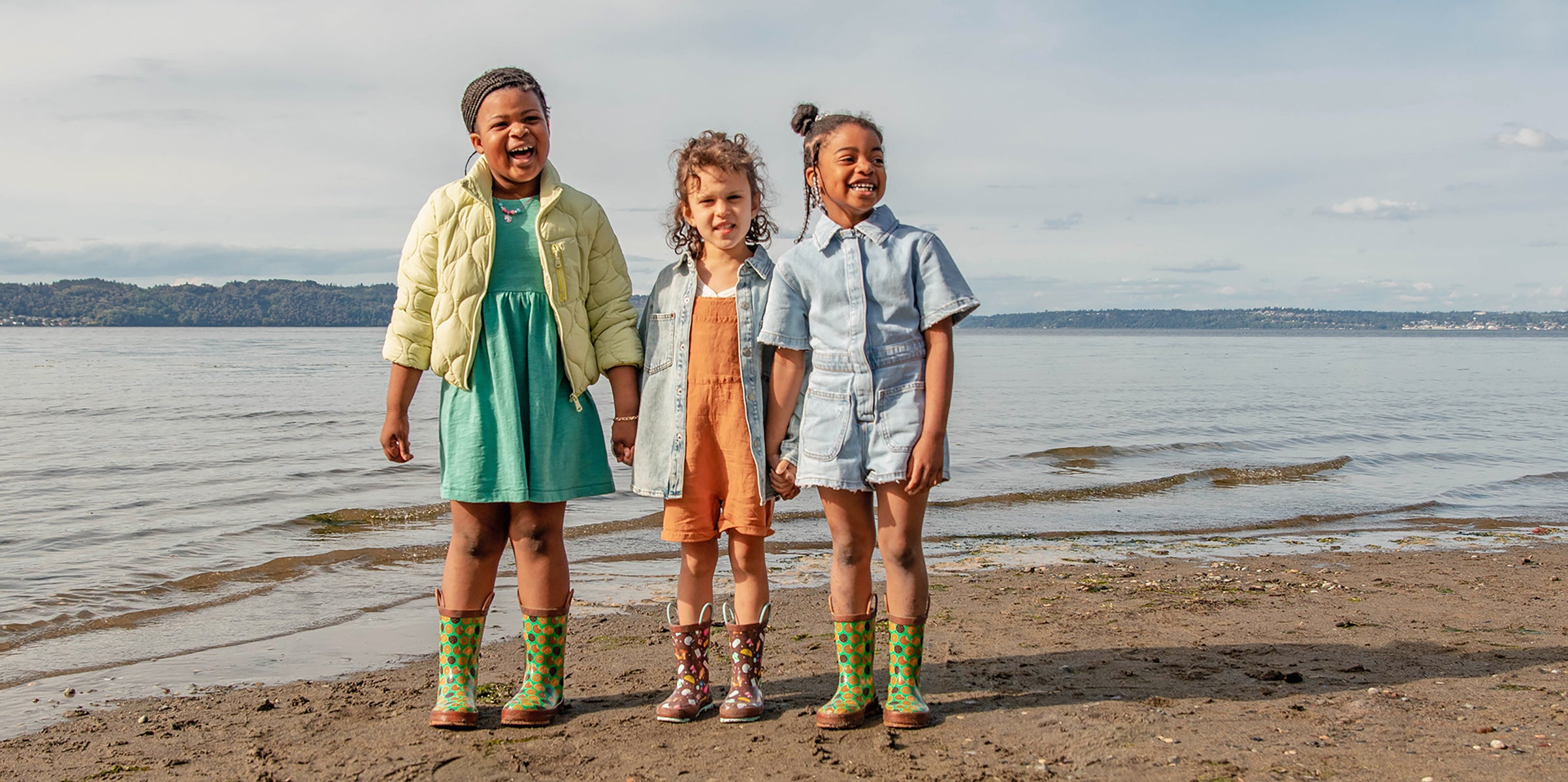 Three smiling girls wearing Western Chief Girl Scouts themed Rain boots on the beach very close to the water.